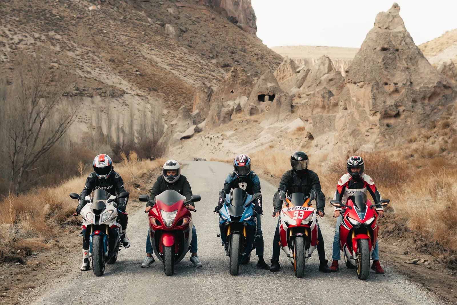 Five riders lined up side by side across an empty road through a rocky desert valley