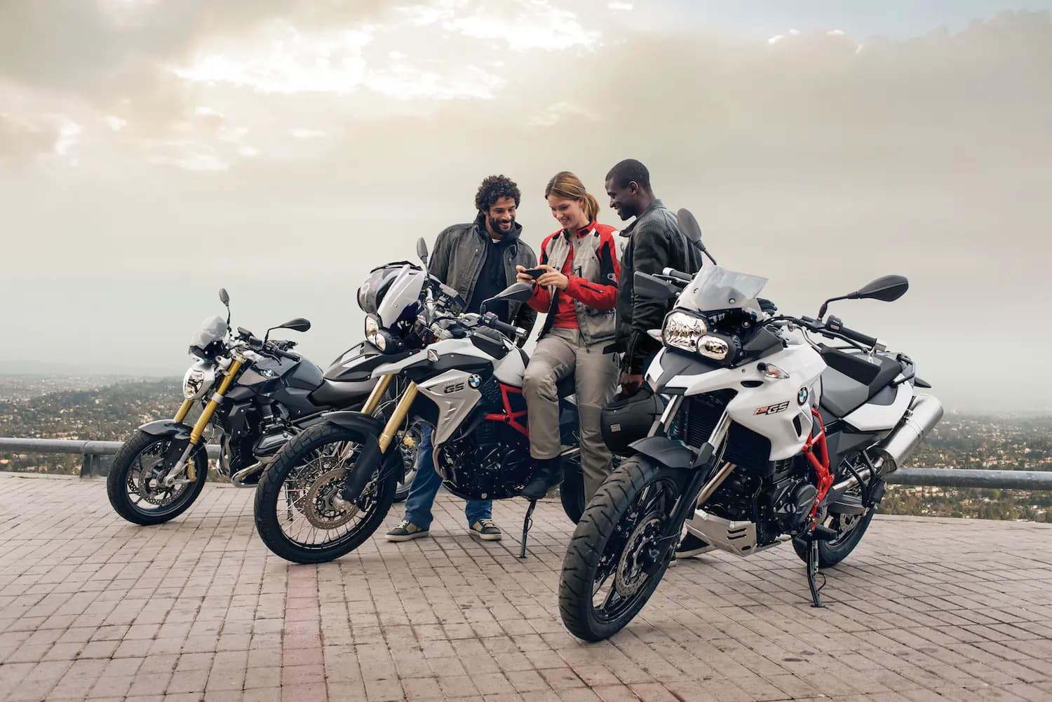 Three riders standing beside their motorcycles at a hilltop viewpoint above the city, looking at a phone together while planning a ride