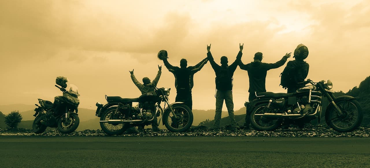 Silhouettes of five riders raising their arms beside their parked motorcycles at a mountain roadside pull-out at sunset