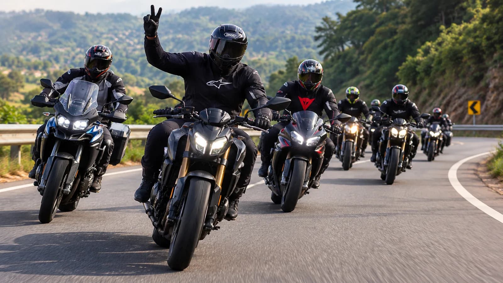 An informal group of riders on mixed motorcycles cruising a winding hillside road, the lead rider raising a two-finger salute